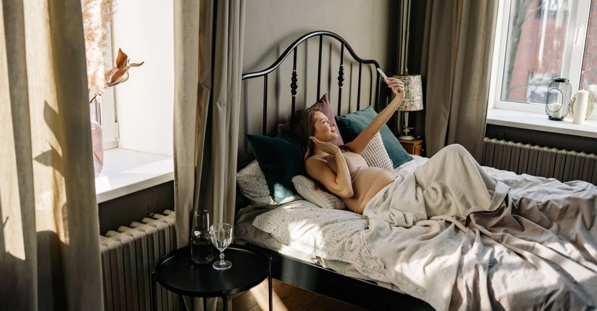 Femme enceinte allongée sur le lit en train de prendre un selfie, profitant d'un matin paisible dans une chambre ensoleillée.