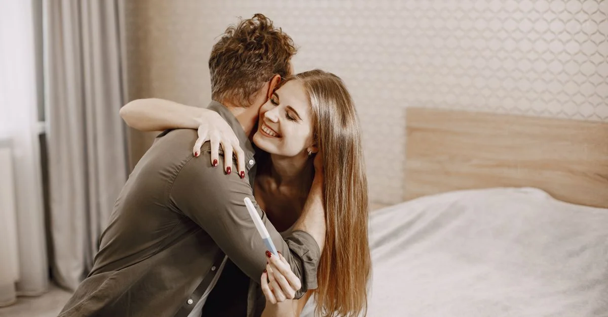 Un couple heureux s'étreignant sur un lit, tenant un test de grossesse positif dans un cadre intime.