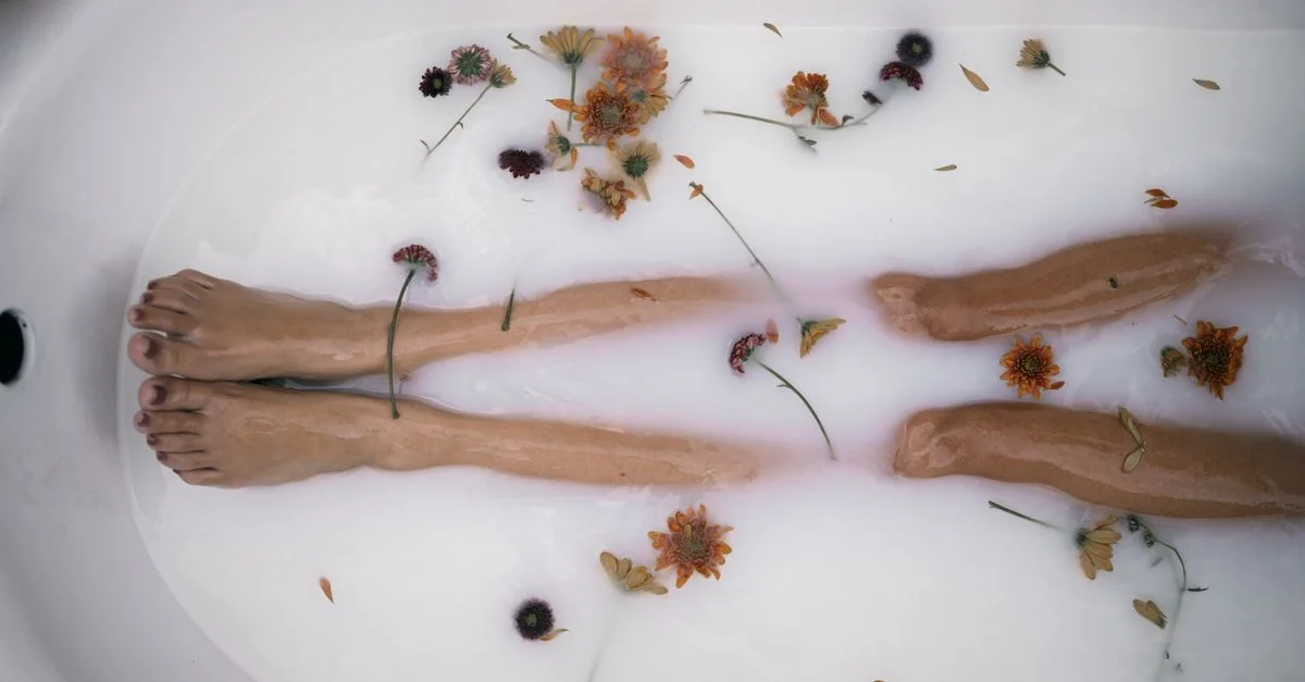 Scène de bain sereine avec des jambes immergées et des fleurs éparpillées, créant une atmosphère paisible.