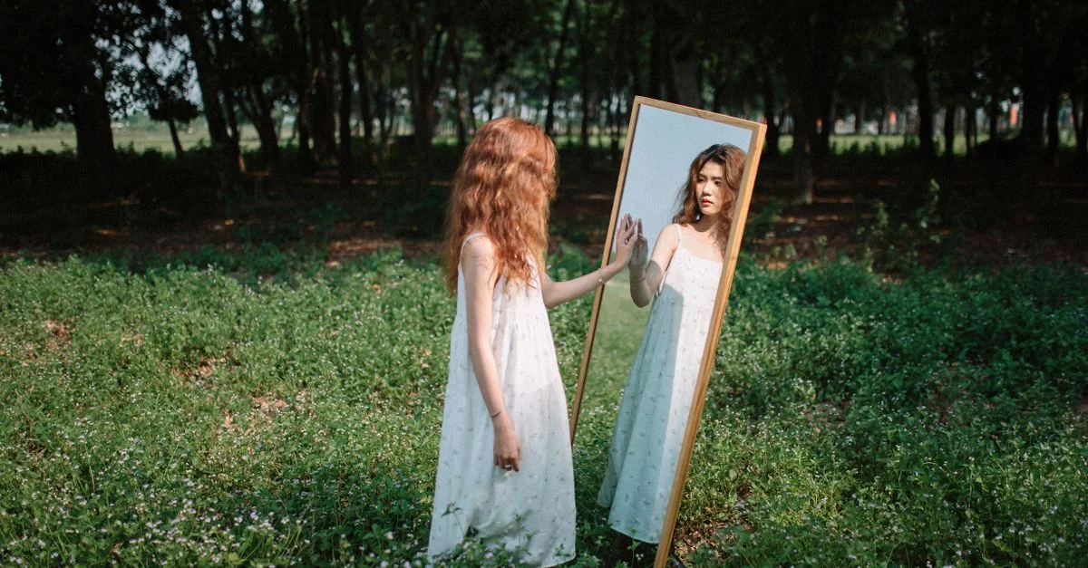 Jeune femme en robe blanche contemplant son reflet dans un miroir au milieu d'un parc verdoyant.