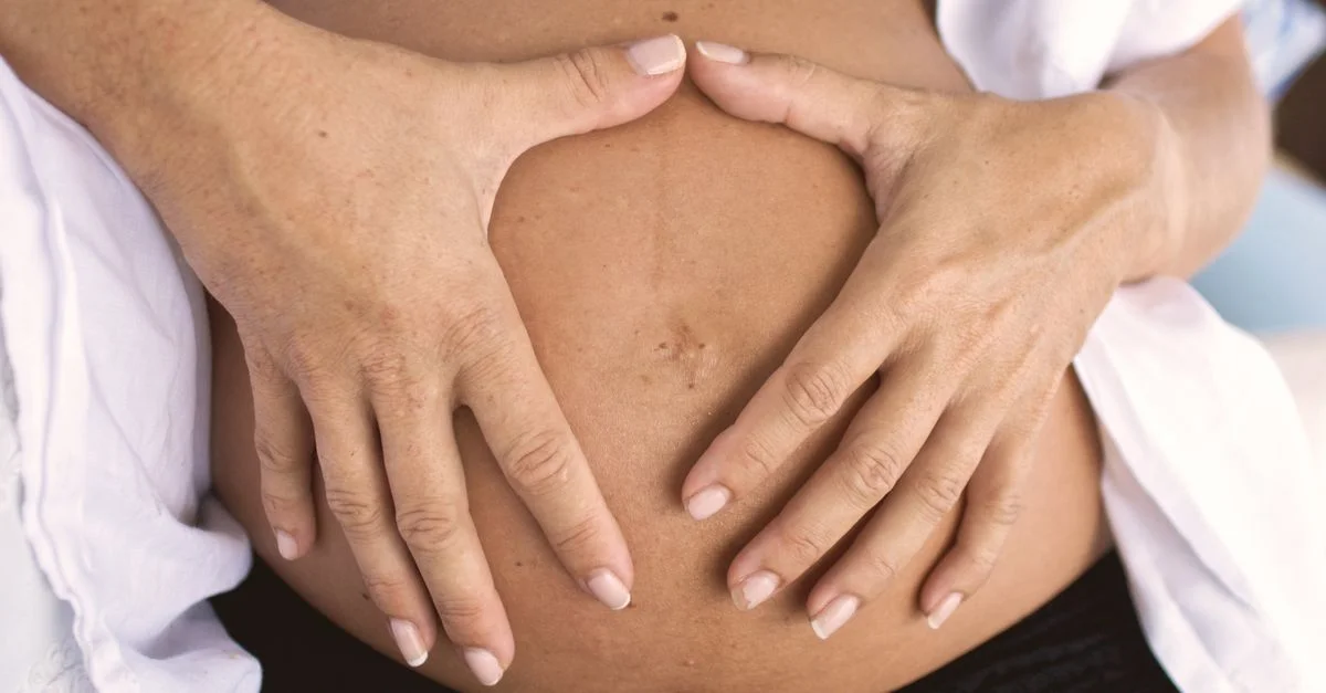 Gros plan sur un ventre de femme enceinte avec des mains formant une forme de cœur, symbolisant l'amour et la maternité.