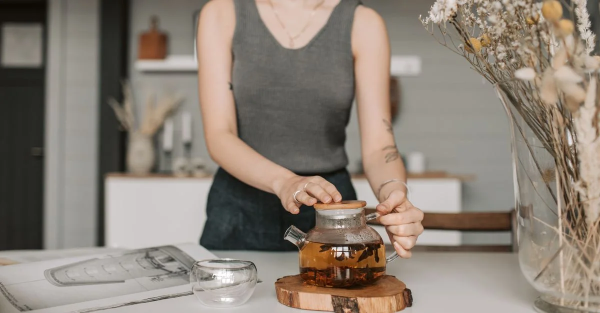 Une femme en débardeur gris prépare une tisane dans une théière en verre sur une table blanche.