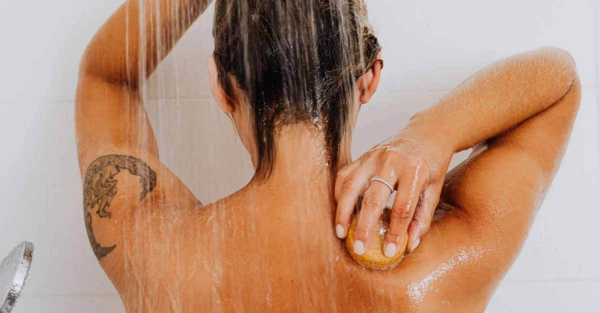 Femme se frottant le dos sous l'eau de la douche, mettant en avant sa routine de soins du corps.