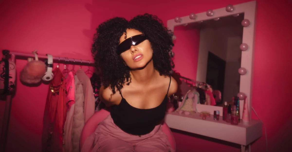 Femme aux cheveux bouclés et lunettes de soleil posant avec style dans une pièce rose vibrante.