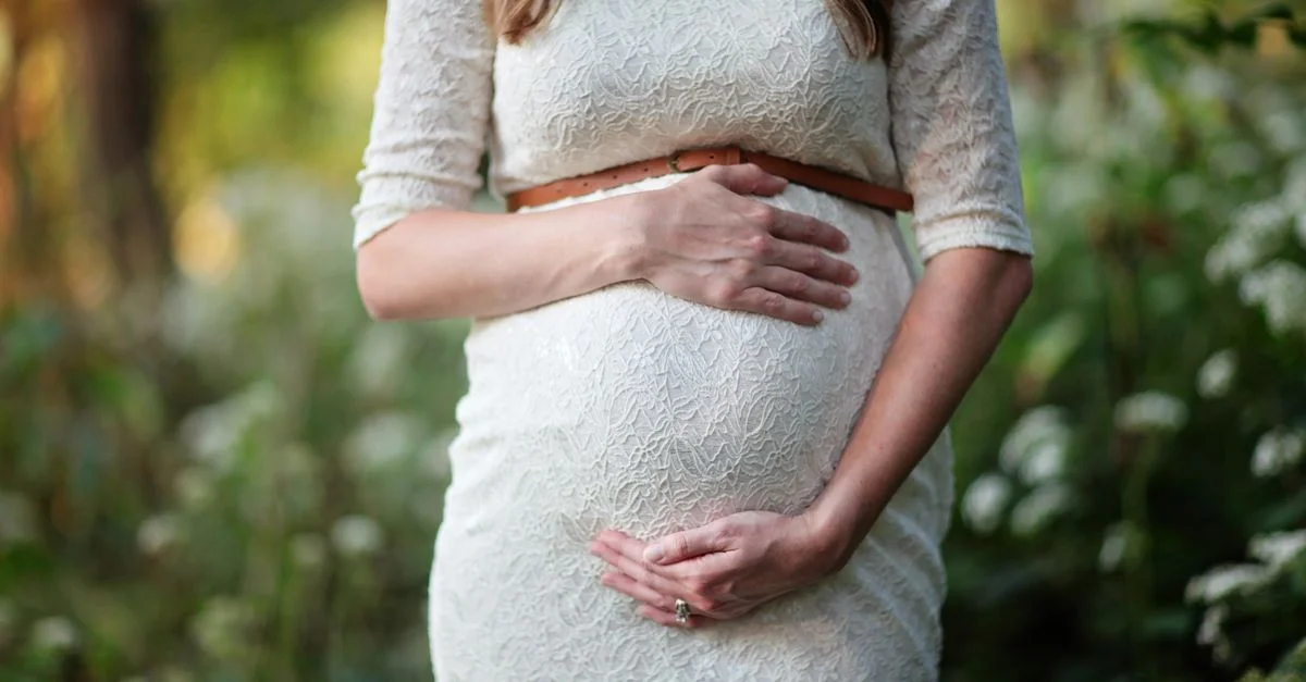 Une femme enceinte en robe en dentelle caressant doucement son ventre dans un cadre extérieur.