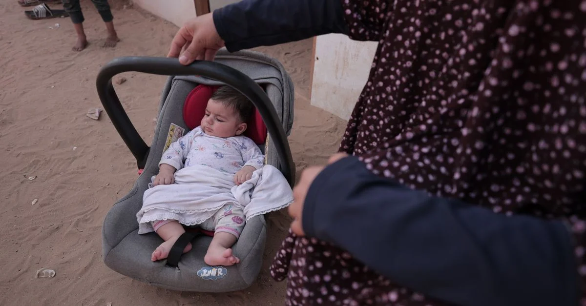 Un bébé dort dans un siège auto à l'extérieur, tenu par une femme vêtue d'une robe à fleurs, créant une scène paisible.