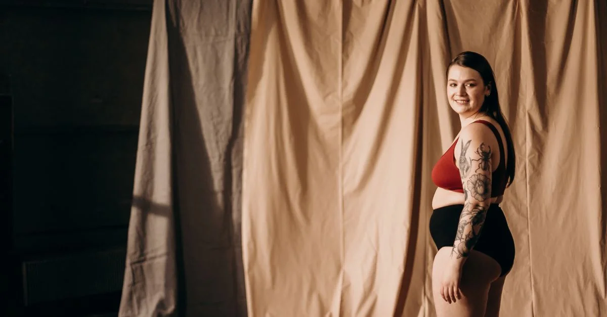 Femme souriante en lingerie avec un tatouage posant avec confiance à l'intérieur. Thème de l'empowerment et de la positivité corporelle.