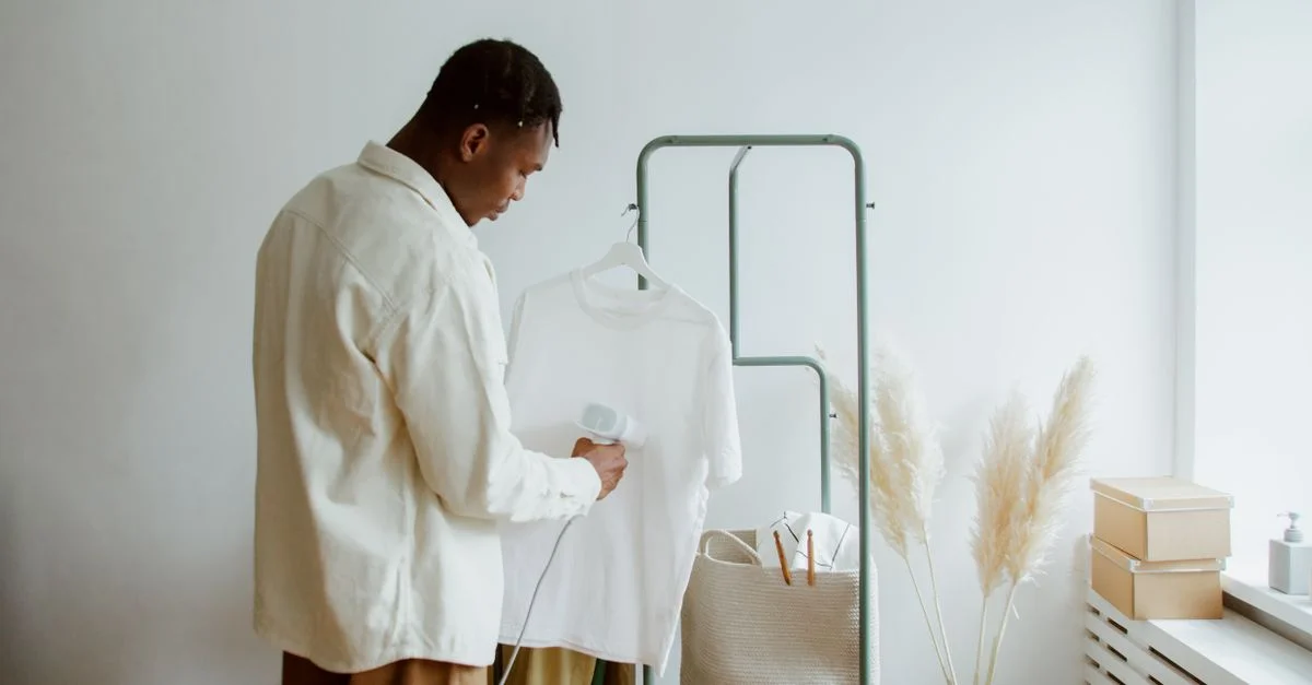 Un homme repasse un t-shirt blanc sur un porte-vêtements à l'intérieur, mettant en avant un style minimaliste.