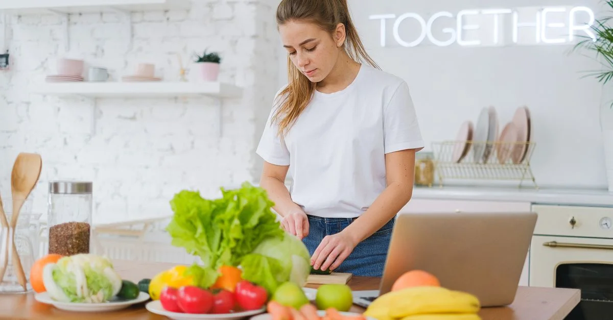 Femme en t-shirt blanc préparant des légumes frais dans une cuisine lumineuse avec un ordinateur portable.