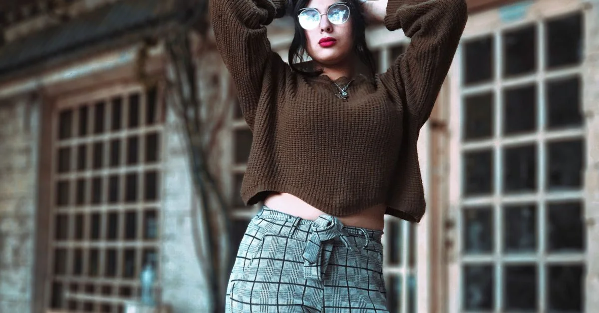 Femme élégante avec des lunettes posant en plein air avec un pull tendance et un pantalon à carreaux.