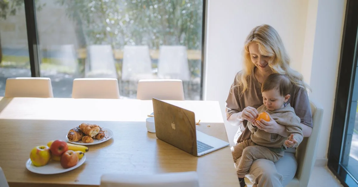 Mère blonde tenant son bébé avec un ordinateur portable à la table à manger, profitant de son petit-déjeuner à l'intérieur.