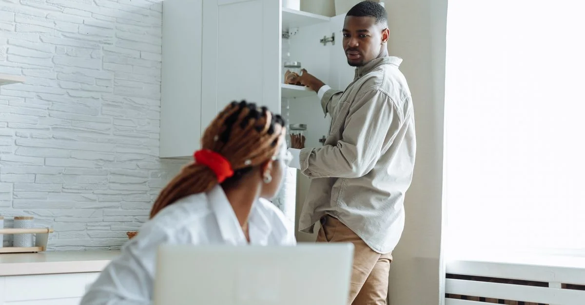 Un couple interagissant dans une cuisine moderne avec un ordinateur portable et un placard ouvert, capturant une scène chaleureuse à la maison.