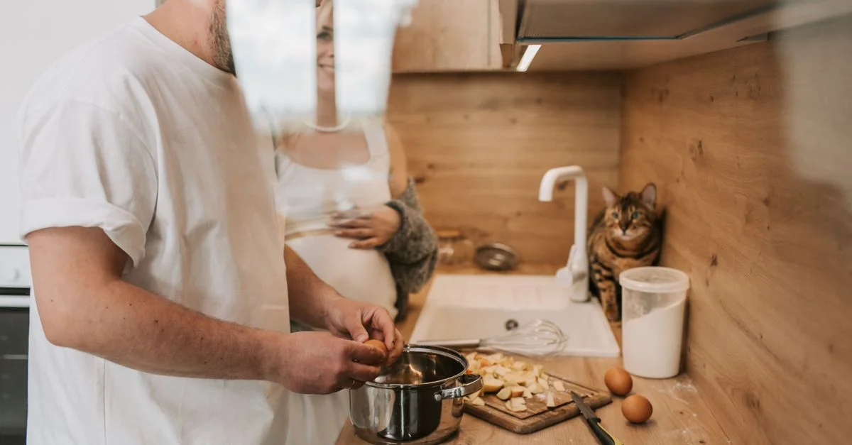 Une femme enceinte et un homme cuisinent ensemble dans une cuisine chaleureuse, accompagnés de leur chat de compagnie.