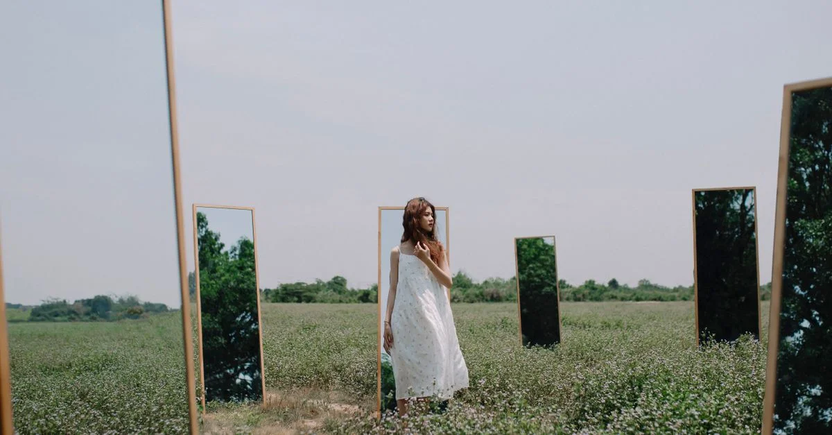 Une femme en robe blanche se tient parmi des miroirs dans un environnement rural, créant une scène surréaliste.