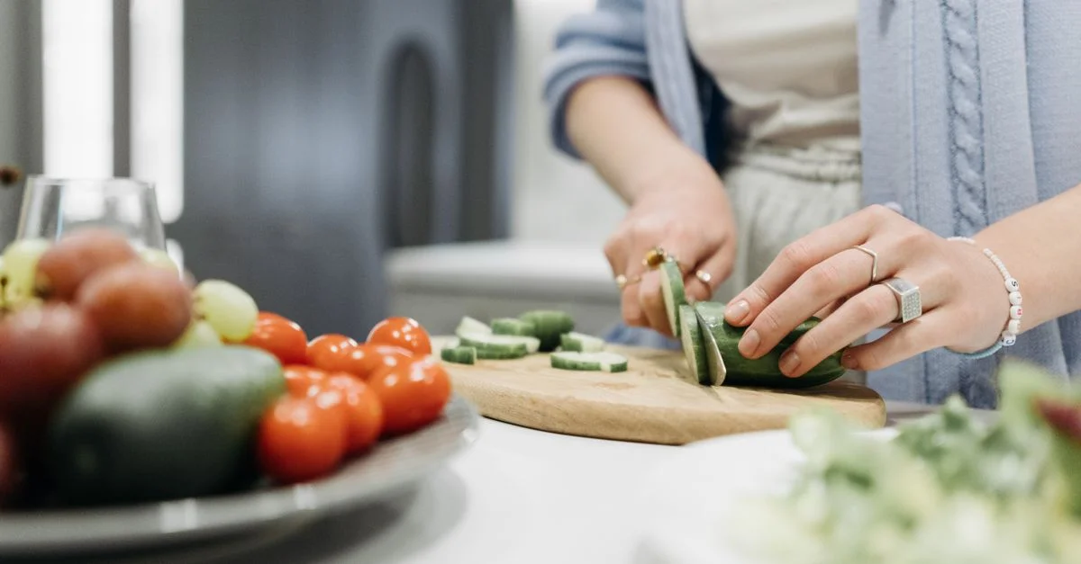 Gros plan sur une femme tranchant des concombres sur une planche en bois entourée de légumes frais.