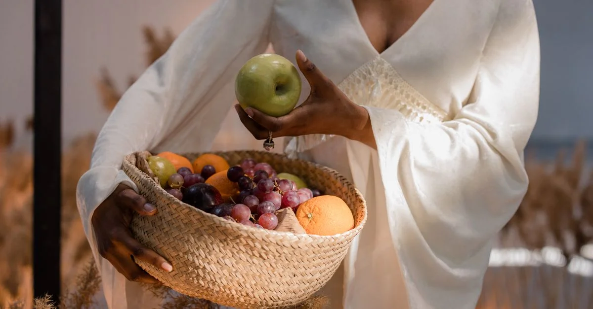 Femme élégante en blanc tenant un panier tissé de fruits frais, mettant en avant un style de vie moderne.