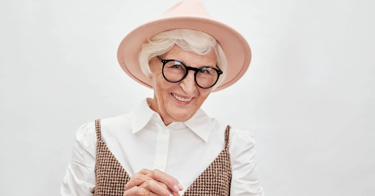 Portrait d'une femme senior aux cheveux blancs et lunettes, souriante tout en portant un chapeau élégant.
