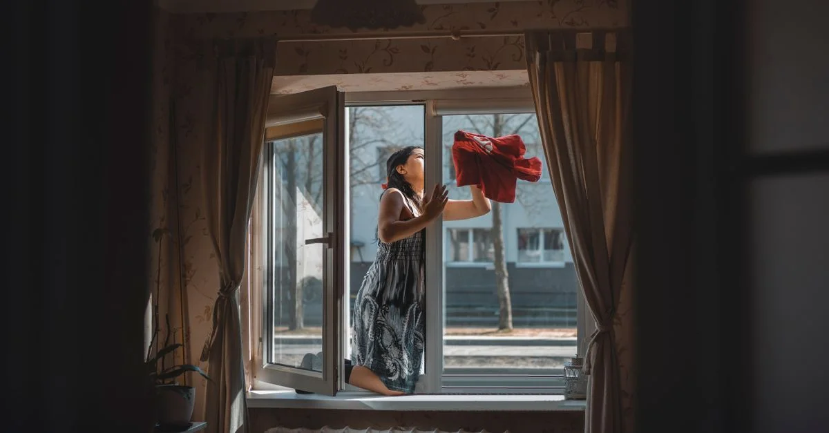 Une femme nettoyant une fenêtre dans un appartement cosy bien conçu avec des rideaux et de la lumière naturelle.